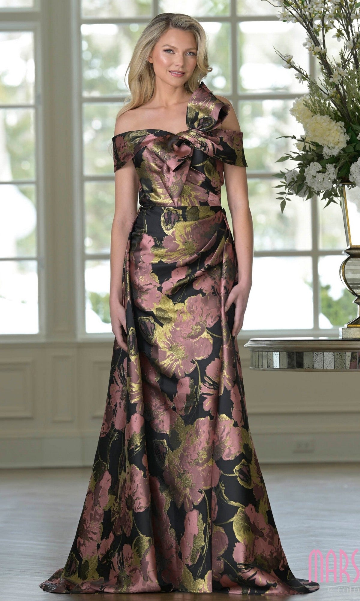 A woman stands indoors near a table with a large flower arrangement, wearing the Marsoni MV1396 long formal dress in Rose Wood Mu.